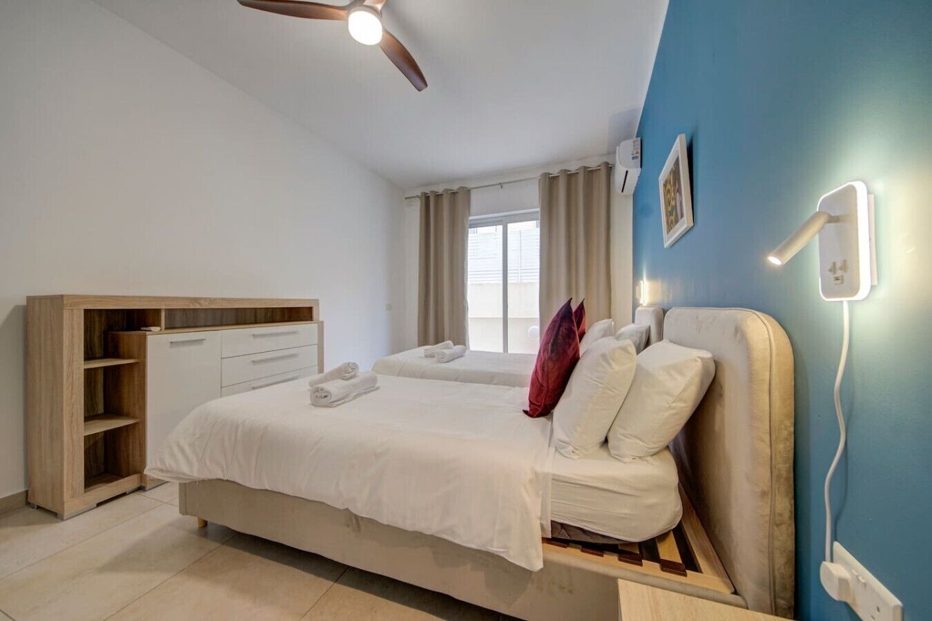 Un dormitorio con cama, mesitas de noche, armario, ventilador de techo, ventana con cortinas y una lámpara de pared.
