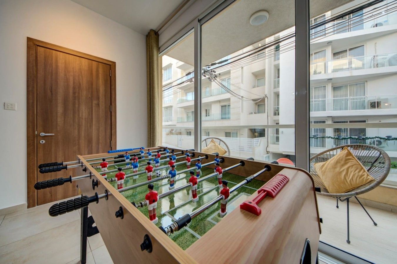 A room with a foosball table, a wicker chair, and a view of a residential building through large windows.