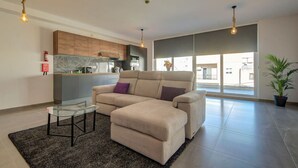 Uma sala de estar moderna com um sofá bege, uma mesa de centro de vidro e uma cozinha ao fundo.