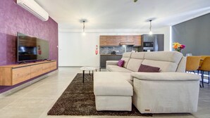Uma sala de estar moderna com um sofá seccional, uma TV de tela plana, uma mesinha de centro e uma cozinha ao fundo.