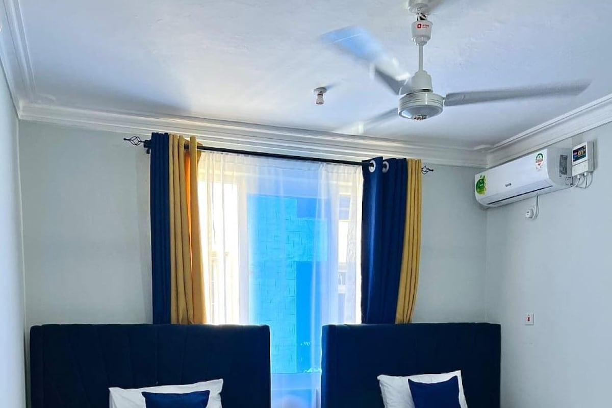 A hotel room with two beds, a ceiling fan, an air conditioning unit, and a window with curtains.