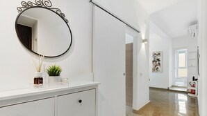 Un couloir avec un mur blanc, un miroir rond, une armoire blanche avec des tiroirs, une plante en pot et une porte donnant sur une autre pièce.