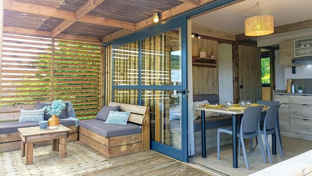 Un rincón al aire libre acogedor con sofá y mesa, una mesa de comedor con sillas y una cocina visible a través de puertas de vidrio.