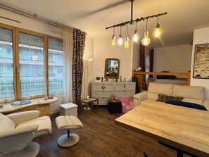 Ein gemütliches Wohnzimmer mit Holzesstisch, einer Couch und einem kleinen Beistelltisch. Es gibt ein Fenster mit gemusterten Vorhängen, einen Spiegel über der Kommode und eine Deckenleuchte.