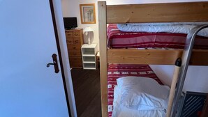 Uma cama beliche com colcha estampada em vermelho e branco, um aparador de madeira, uma televisão e um abajur em um quarto com paredes brancas e teto inclinado.