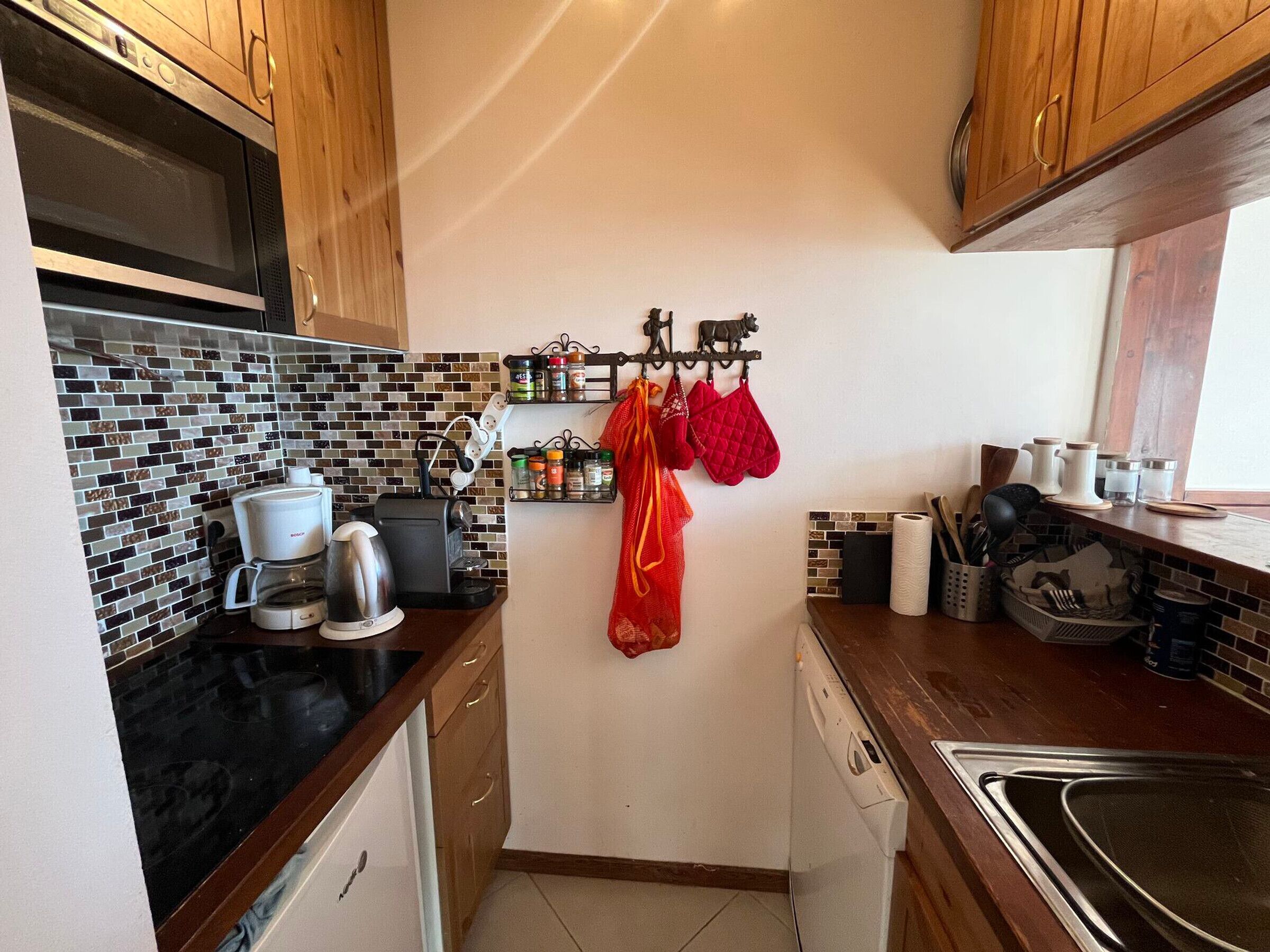 Een kleine keuken met een zwart aanrecht, een koffiezetapparaat, een waterkoker en een magnetron. Er zijn houten kastjes en een betegelde spatwand.