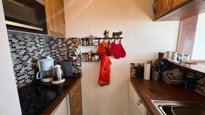 Uma cozinha compacta com bancada preta, cafeteira, chaleira e micro-ondas. Possui armários de madeira e painel de azulejos.