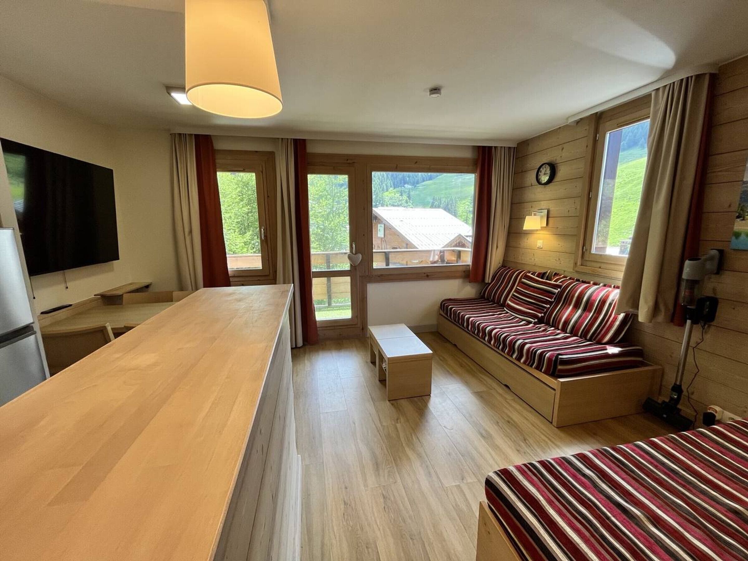Quarto moderno com mesa de jantar de madeira, um sofá, uma TV e janelas amplas com cortinas.