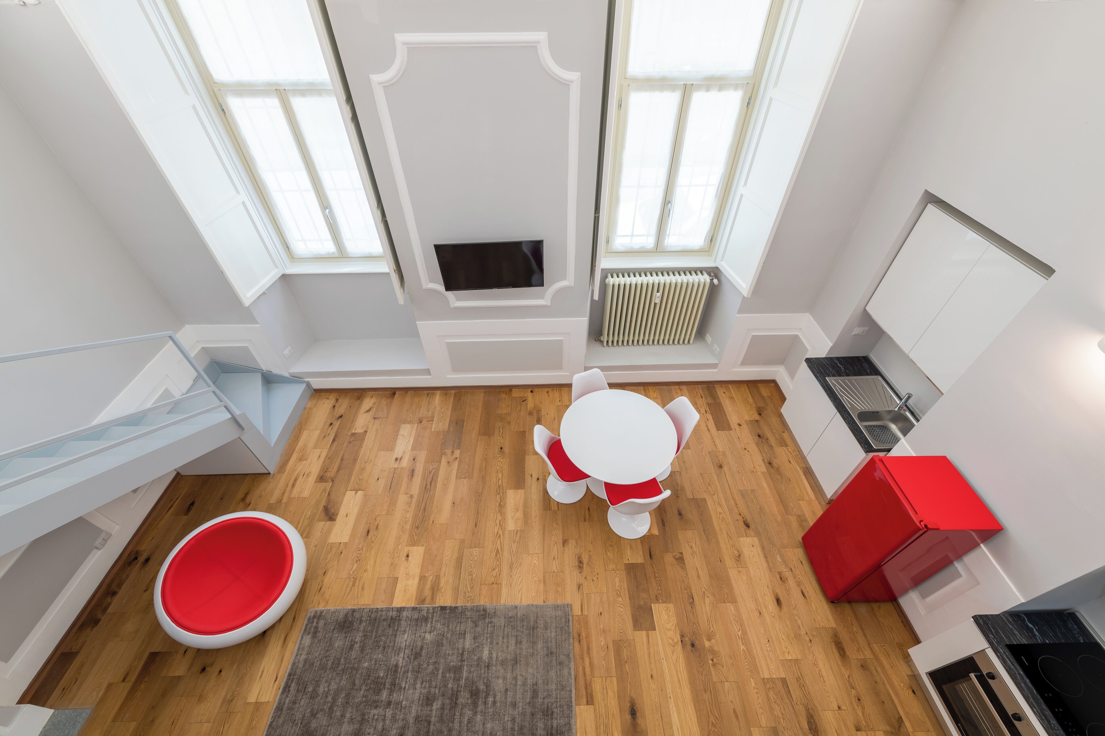 A modern kitchen with a wooden floor, white cabinetry, a red refrigerator, and a built-in oven.
