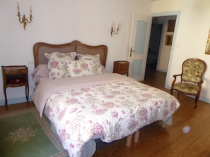 A bedroom with a large bed, floral bedding, two bedside tables, a wooden chair, and a door leading to another room. - Maison Marius—pets allowed—1,600 m² fenced-in garden—heated pool (Le Buisson-de-Cadouin)
