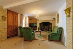 A living room with a fireplace, green upholstered chairs, a television, and a chandelier. - Maison Marius—pets allowed—1,600 m² fenced-in garden—heated pool (Le Buisson-de-Cadouin)