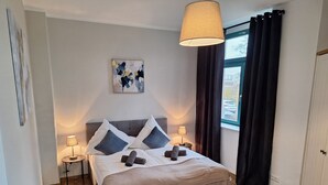Ein modernes Schlafzimmer mit einem Bett, zwei Nachttischlampen, einem Fenster mit Vorhängen, einem Bild an der Wand und einem Holzboden.