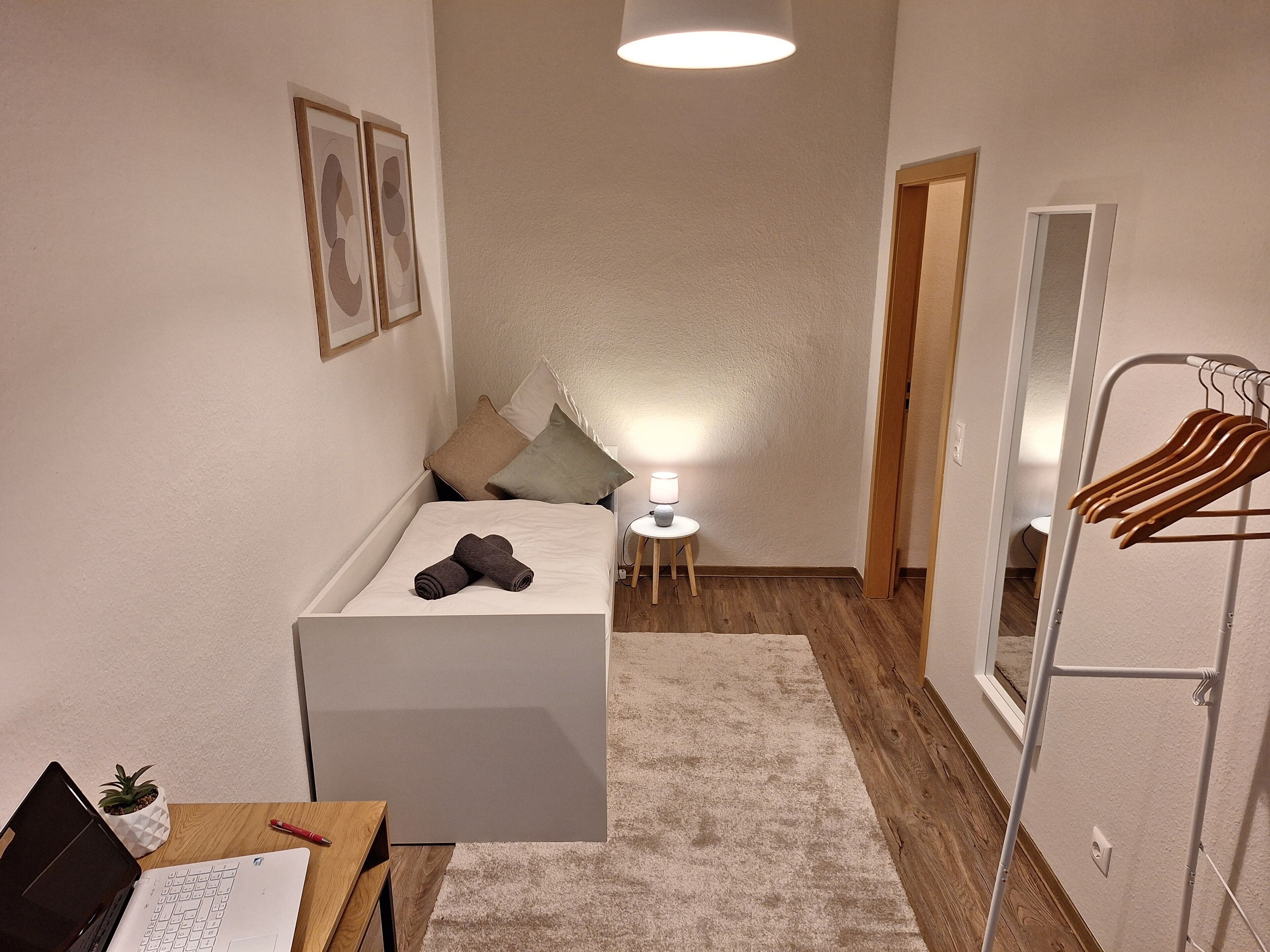 Une chambre à coucher de taille réduite, comprenant un lit, une table de chevet, une lampe et un portant à vêtements.