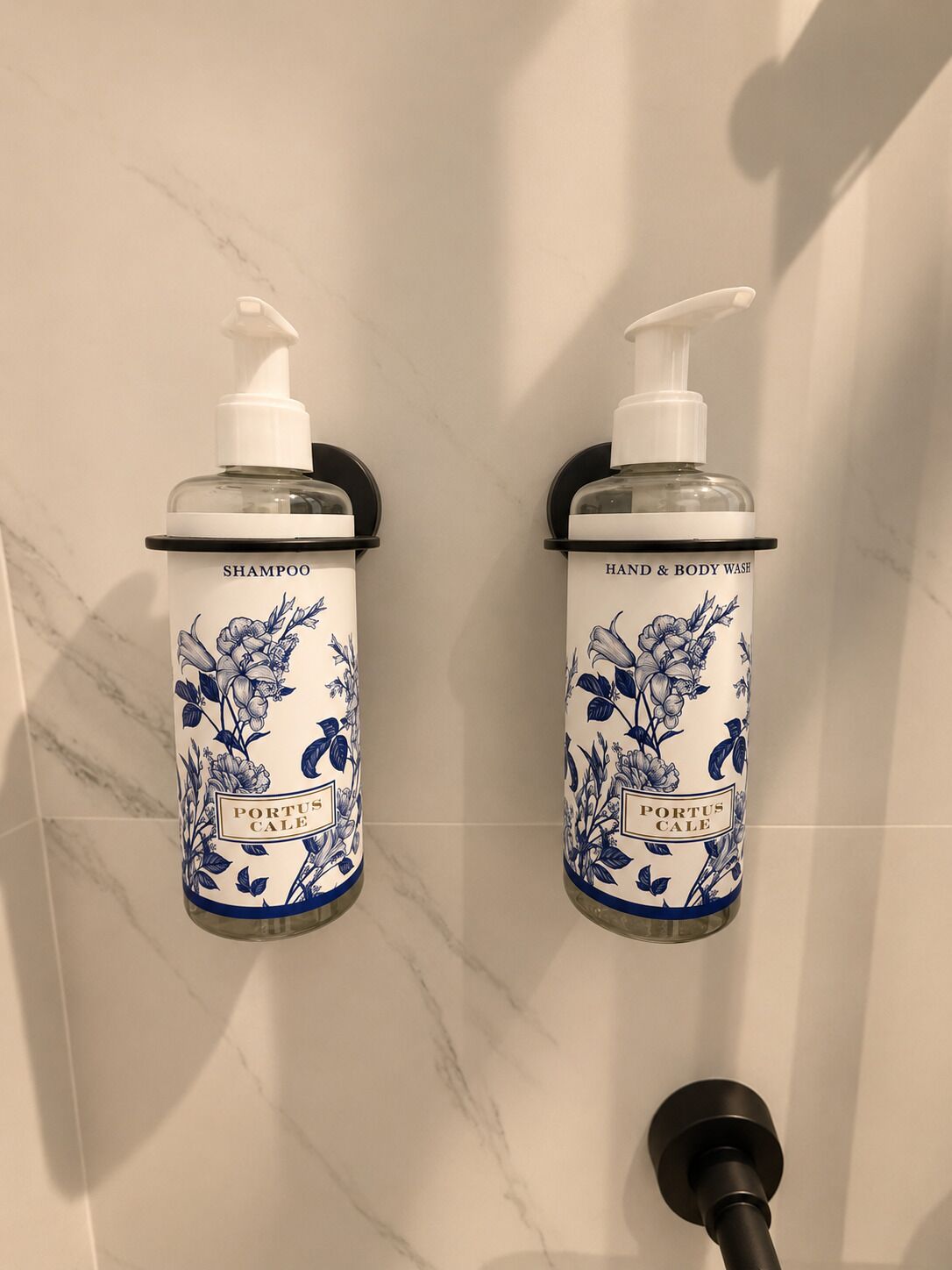 Zwei Toilettenflaschen mit Pumpspender, beschriftet mit 'SHAMPOO' und 'HAND & BODY WASH', beide mit 'PORTUS CALE'-Branding, an einer Wand mit Marmormuster befestigt.