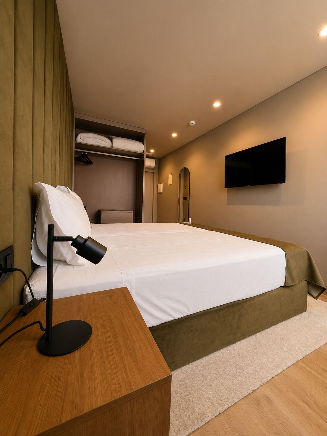 A hotel room with a bed, a desk with a lamp, a TV on the wall, and a wardrobe.