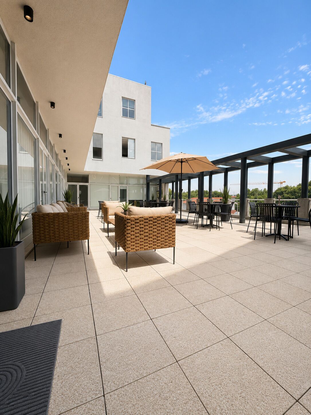 Un espace extérieur aménagé avec des meubles en osier et des parasols, un bâtiment moderne avec de grandes fenêtres en arrière-plan, et un ciel dégagé.
