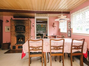 A dining room with a brick fireplace, wooden chairs, a table with a lace tablecloth, and a window with lace curtains.