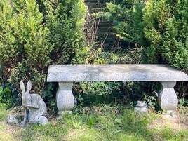 Un banco de piedra con una escultura de conejo y otra escultura más pequeña a su lado, rodeado de follaje.