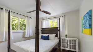 A four-poster bed with a white bedspread and a blue pillow, a white nightstand with a plant, and a ceiling fan. - "Waimea Vista" | Oceanfront, Walkable to Beaches (Haleiwa)