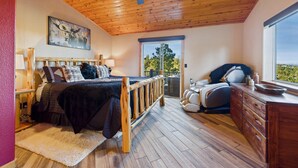 A bedroom with a wooden ceiling fan, a bed with a dark blanket, a massage chair, and a view of the outdoors through a window. - Salty Pines - Scenic Log Cabin w/ Hot Tub, Sierra Blanca Views & Pet-Friendly (Ruidoso)
