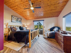 Un dormitorio con ventilador de techo de madera, una cama con manta oscura, una silla de masaje y vistas al exterior a través de una ventana.