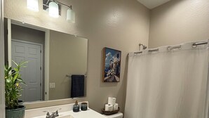 Une salle de bain avec un lavabo blanc, un miroir et des toilettes. On y trouve un rideau de douche, une plante en pot et un tableau accroché au mur.
