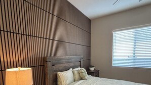 Une chambre avec un mur recouvert de lambris en bois, un lit avec une couvre-lit à rayures, une lampe de chevet, une table de chevet avec des tiroirs, un ventilateur de plafond et une fenêtre avec des stores.