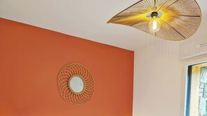 Een net opgemaakt bed met een wit hoofdbord en kussens, een klein krukje, een wandspiegel en een bijzondere plafondlamp.