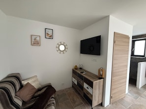 A small, modern living room with a sofa, a TV, a wooden cabinet, and a door leading to another room. - Chez Maud et Fab (Dinozé)