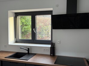 A modern kitchen with a black countertop, a sink, a window with a view of greenery, and a built-in stove. - Chez Maud et Fab (Dinozé)