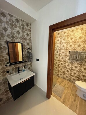 Une salle de bain avec un mur orné d’un motif géométrique, un lavabo blanc avec des robinets noirs, un miroir et une porte en bois donnant sur une autre pièce.