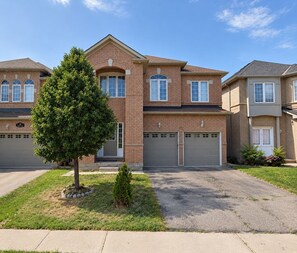 A two-story brick house with a garage, a small tree in the front yard, and a clear sky. - Spacious Private King Bedroom w/Workspace, TV, Mini Fridge, 2.5 Bath—Shared Home (Brampton)