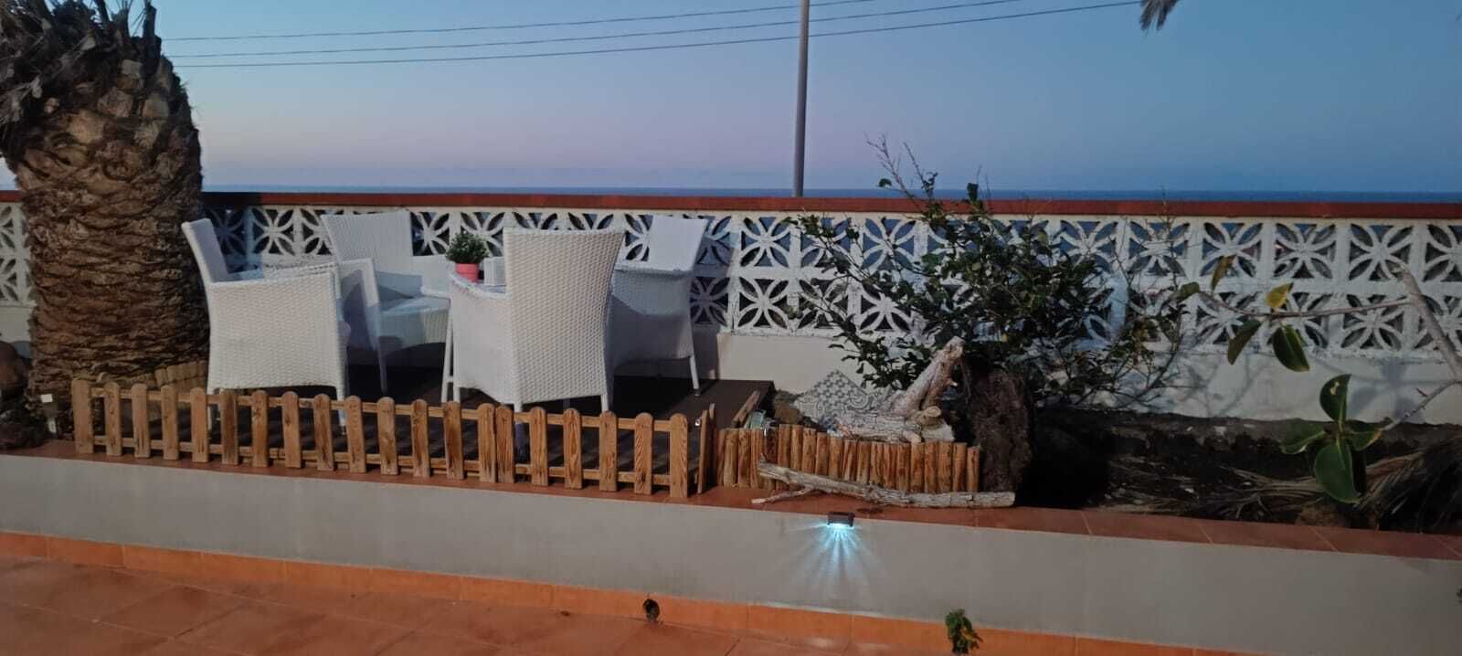 Een terras met rieten stoelen en een kleine tafel, een houten hek en een palmboom.