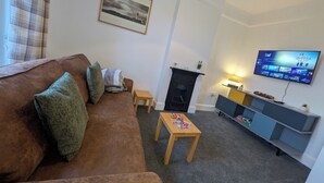 A living room with a brown sofa, a small wooden table with a board game, a fireplace, a television mounted on the wall, and a framed picture above the fireplace. - digby house flat 1 (West Mersea)