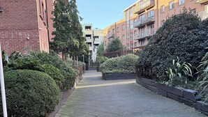 A paved pathway flanked by brick buildings with balconies and dense shrubbery. - Spacious Apartment on a quiet street 1 min from Smithfield Square. (Dublin 7)