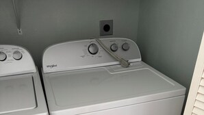 Vista frontal de una unidad de lavadora y secadora Whirlpool blanca, con perillas de control y un agarrador en la parte delantera, ubicada frente a una pared de color claro con piso de baldosas.