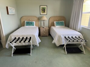 A room with two beds, a small nightstand, a lamp, a framed picture, and a window with curtains. - Little Cayman Time - CC 18 - Beachfront 2-bedroom Condo w/ 2 pools and Jacuzzi (Blossom Village)
