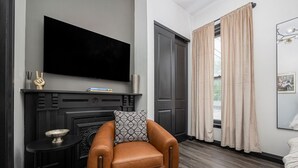 A modern hotel room with a leather armchair, a flat-screen TV mounted above a fireplace, and a large window with curtains. - German Village Gem - Pets Welcome (Columbus)