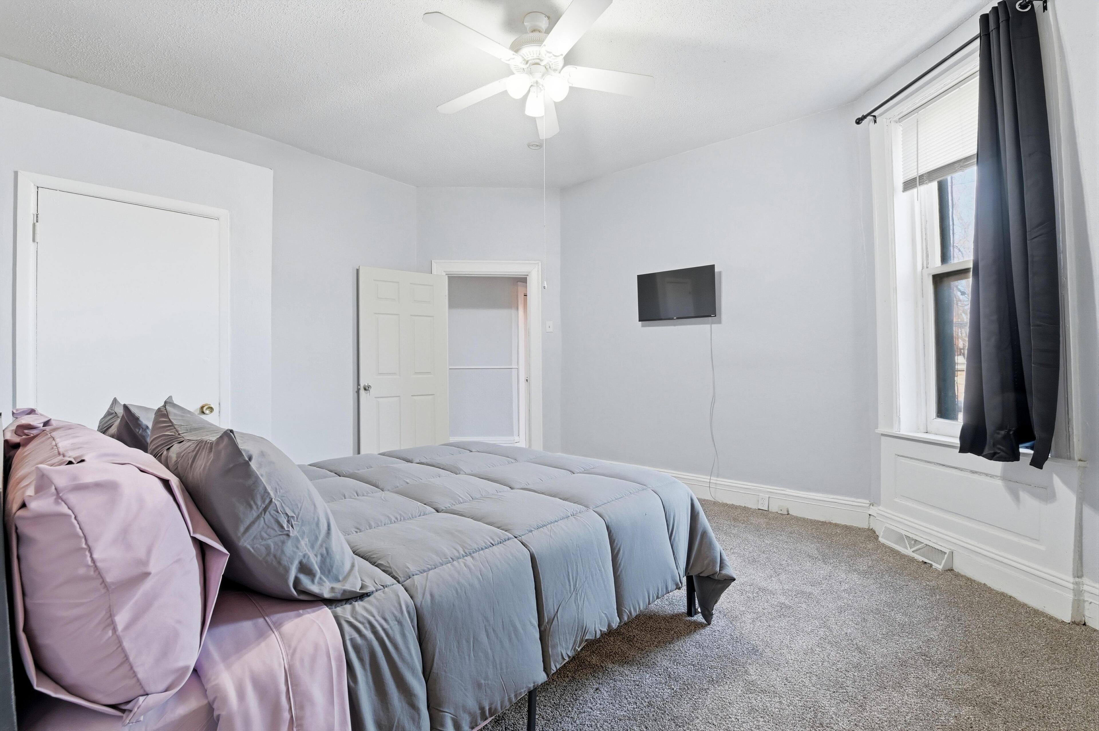 Un dormitorio con cama, ventilador de techo, televisor y ventana con cortinas.