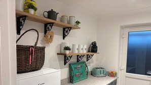 Una cocina con un refrigerador blanco, un tostador y una cesta sobre la encimera. Hay estantes arriba con varios utensilios de cocina.