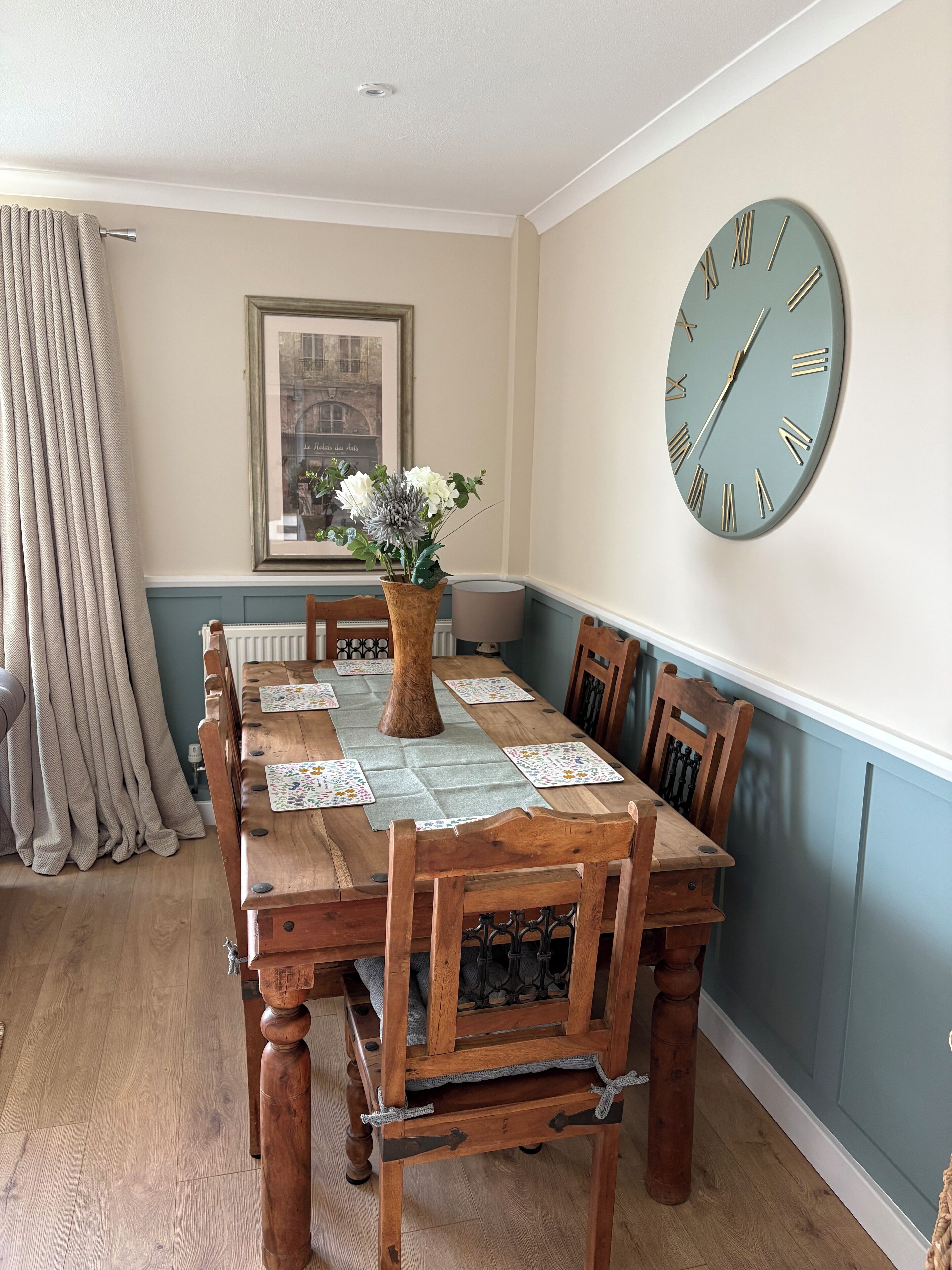 A dining room with a wooden table set for four, a vase of flowers, and a large wall clock.