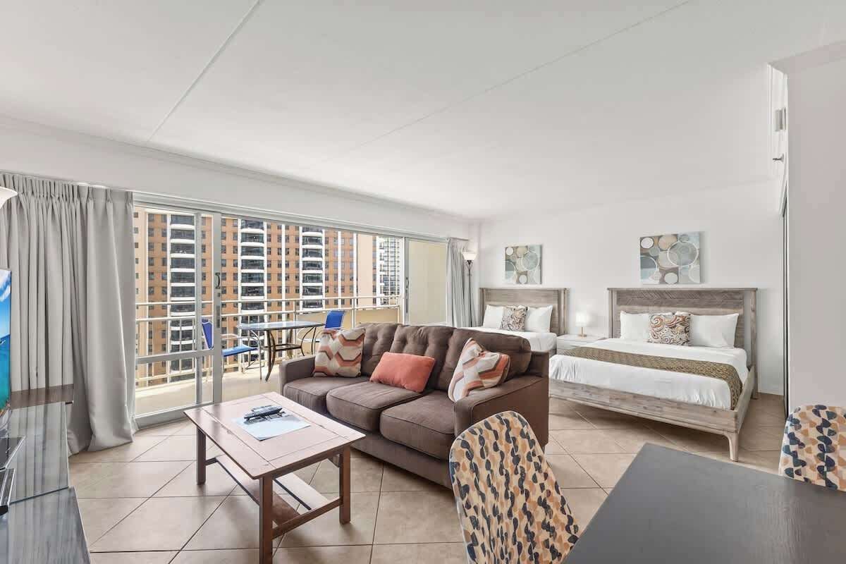 A modern hotel room with a sofa, a bed, a desk, and a balcony with a view of high-rise buildings.