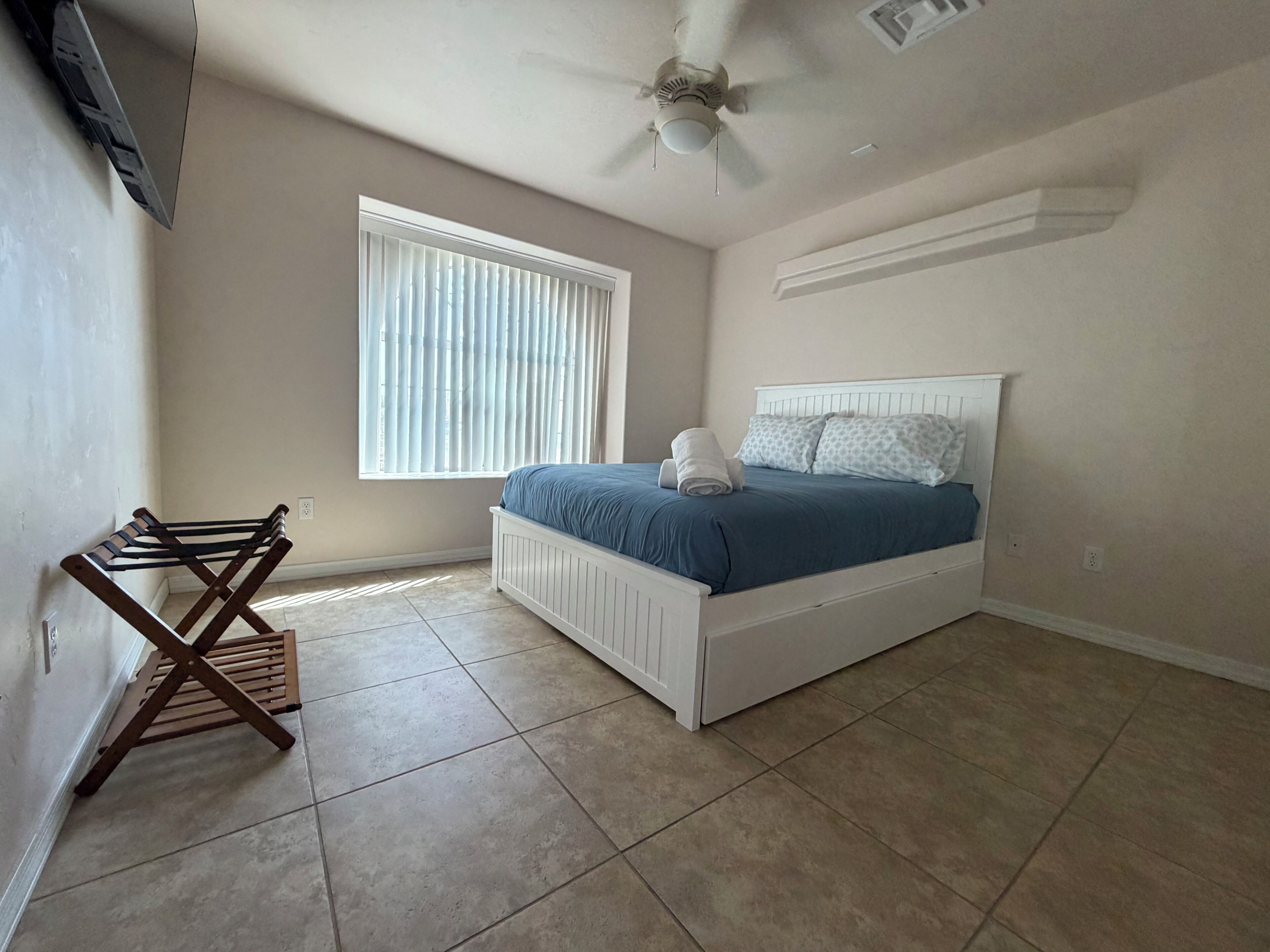A bedroom with a bed, a ceiling fan, a window with blinds, and a wooden chair.
