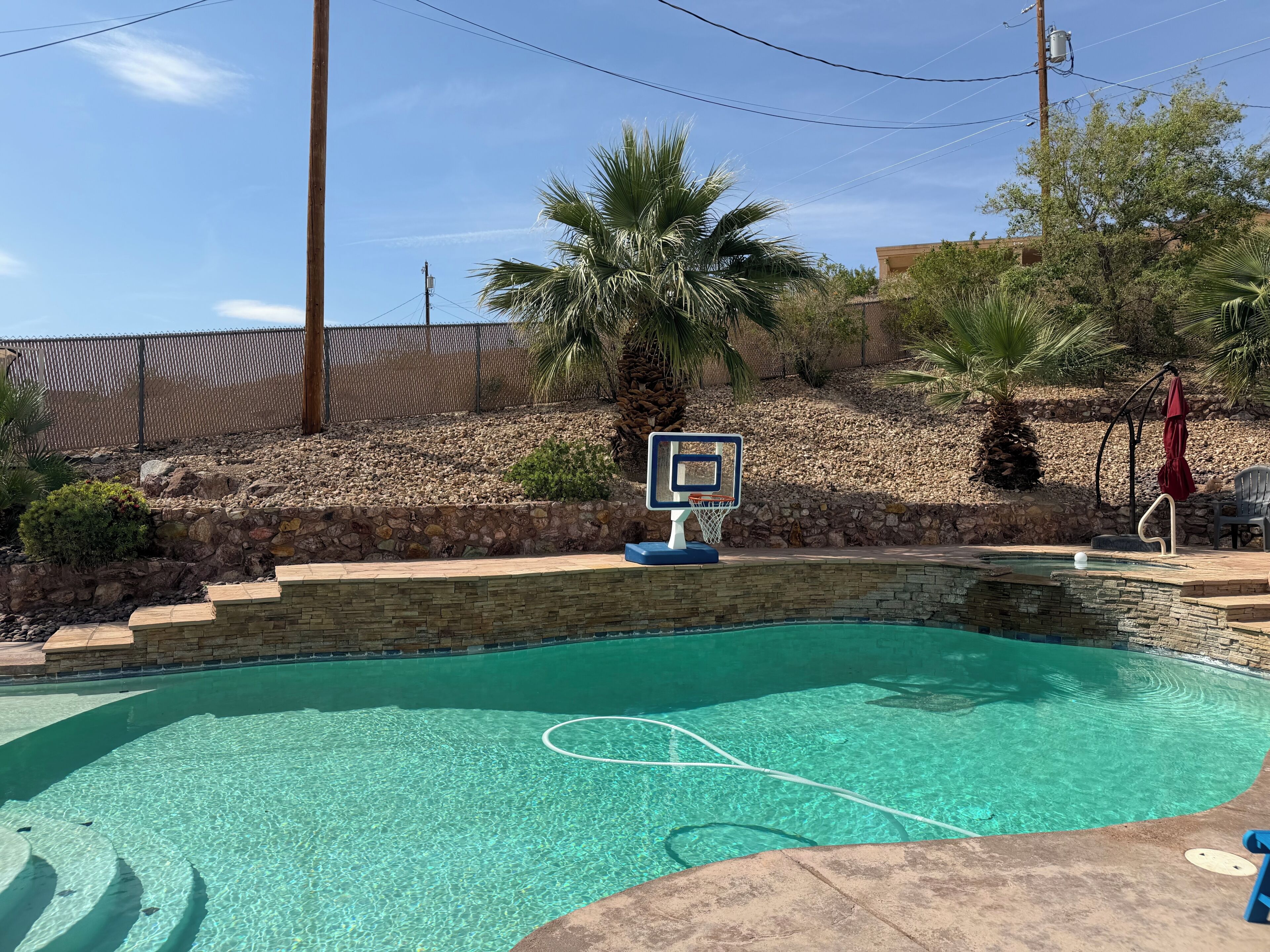 Une piscine dotée d’un panier de basket, entourée d’un mur de pierre et de palmiers.