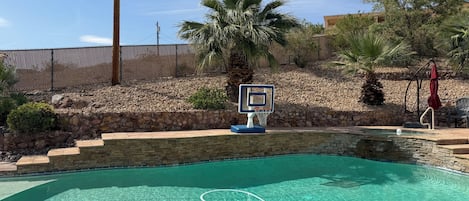 Une piscine dotée d’un panier de basket, entourée d’un mur de pierre et de palmiers.
