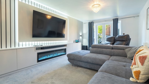 Un salon moderne doté d’un grand téléviseur à écran plat fixé à un mur blanc, d’un canapé d’angle gris et d’une cheminée avec un feu illuminé de bleu.