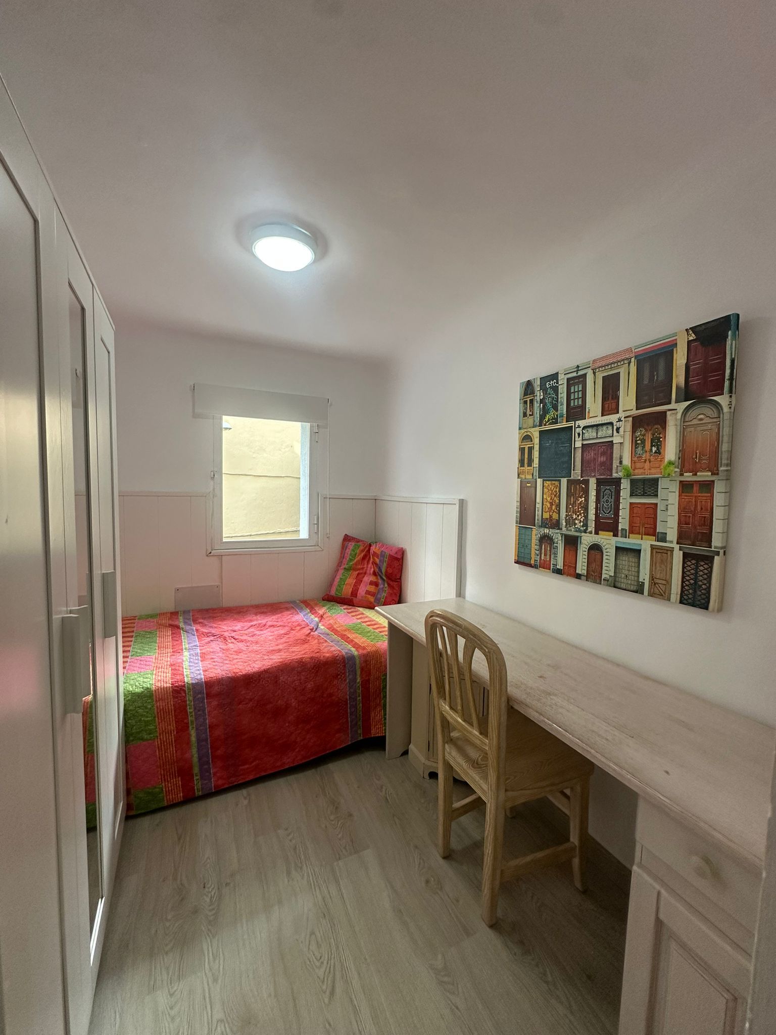 Une chambre à coucher comprenant un lit, un bureau, une chaise, une fenêtre et un mur orné d’une œuvre d’art colorée.