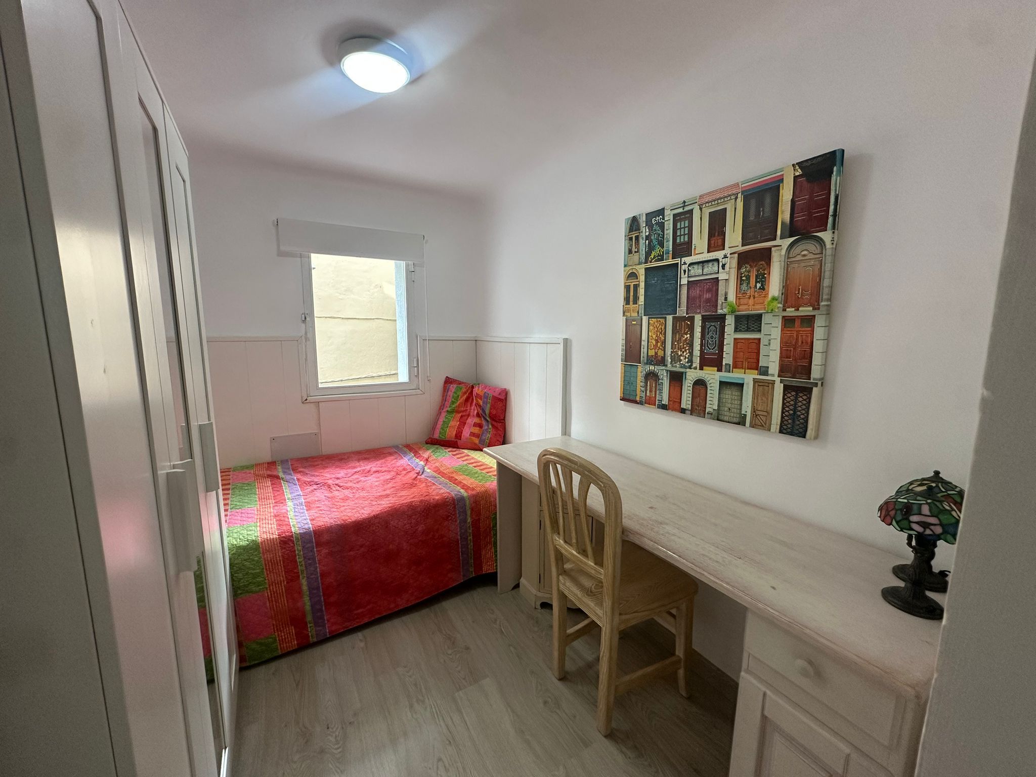 A bedroom with a bed, a desk, a chair, a window, and a ceiling light.