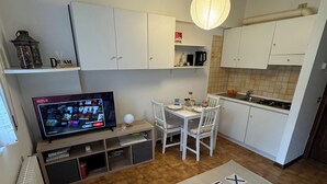 A modern kitchen with white cabinets, a dining area with a round table and chairs, a TV displaying a streaming service, and a decorative rug with a clock pattern.