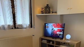 A hotel room with a bed, a television displaying a show, a shelf with decorative items, and a window with curtains.
